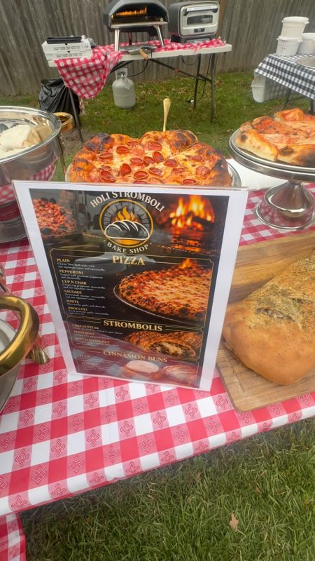 Backyard catering setup with pizza, stromboli, and cinnamon buns displayed on checkered tablecloths next to a menu board