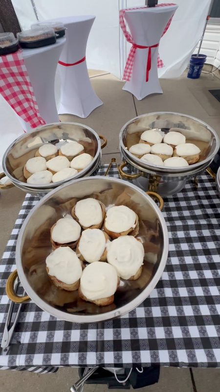 Video still of trays of frosted cinnamon buns in serving bowls at an outdoor catering event